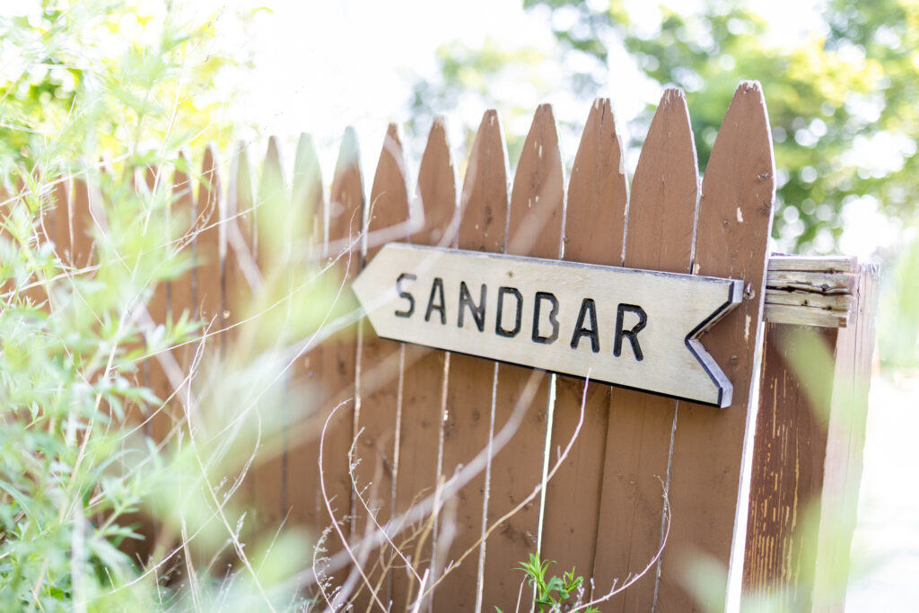 Rustic wooden sandbar directional sign on weathered brown picket fence surrounded by summer foliage